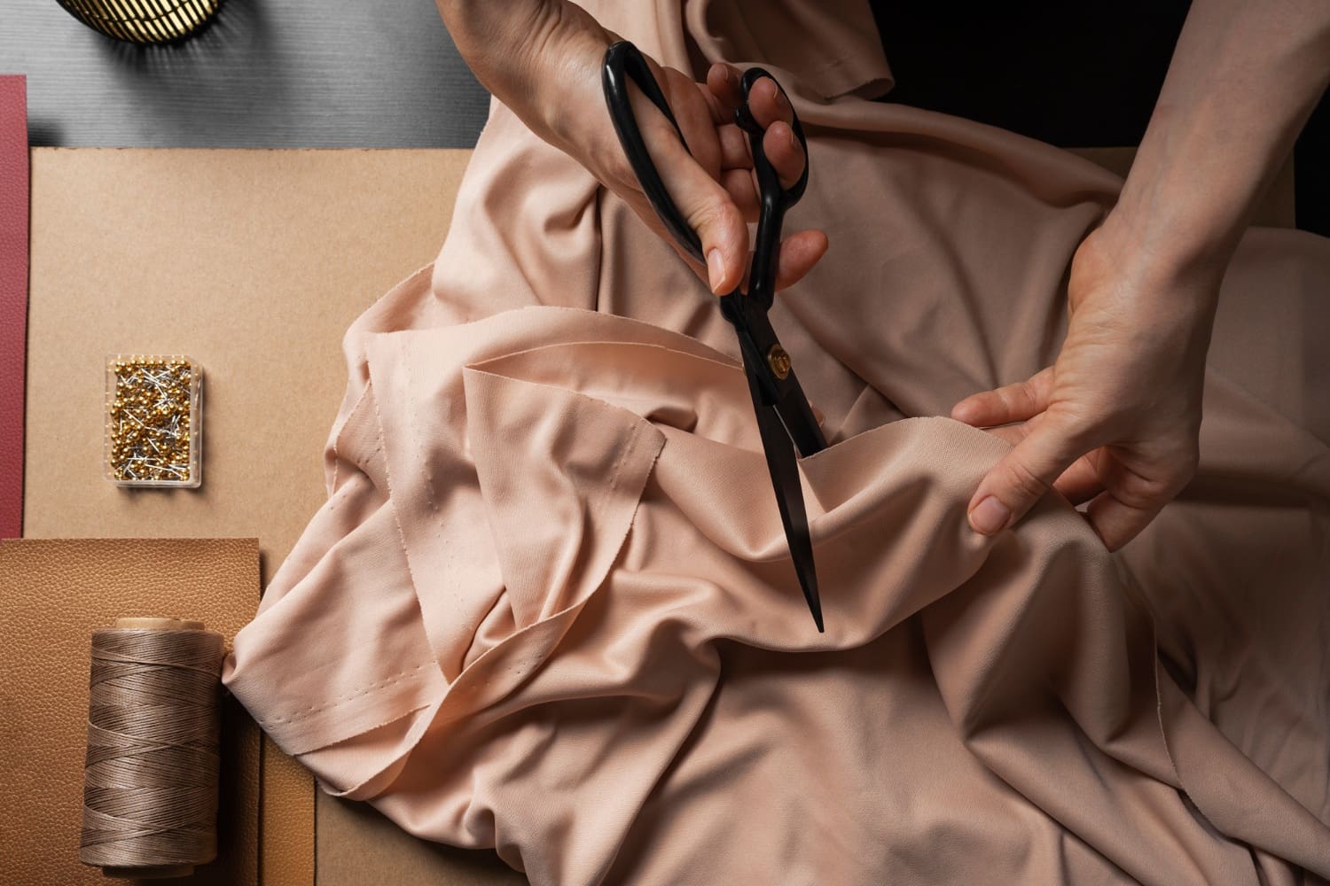 Hands cutting light pink fabric with scissors on a table, surrounded by sewing supplies including thread, fabric rolls, and a case of pins.