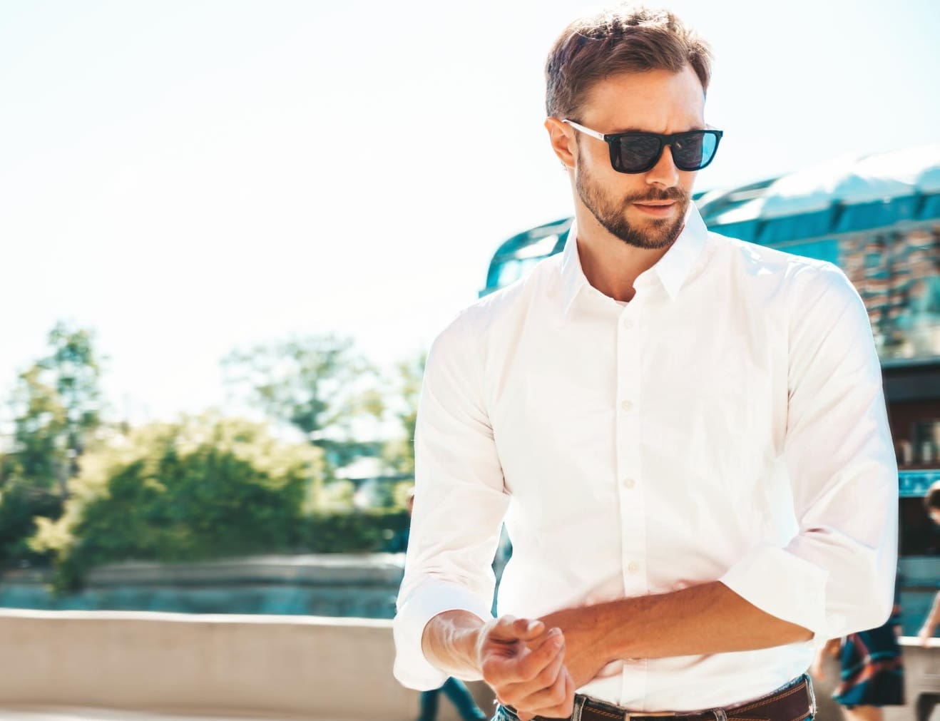 A man wearing sunglasses and a white shirt stands outdoors on a sunny day, with blurred trees and buildings in the background.