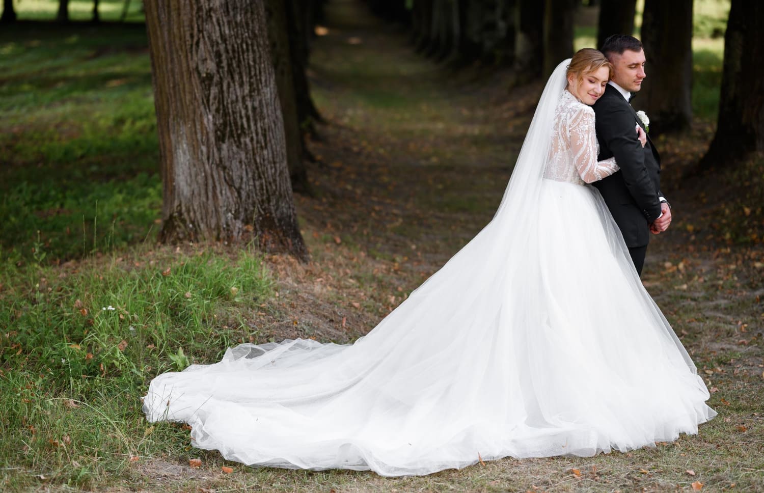 Bride in a long white gown and veil embraces groom from behind in a wooded outdoor setting, both standing on grass near tall trees.