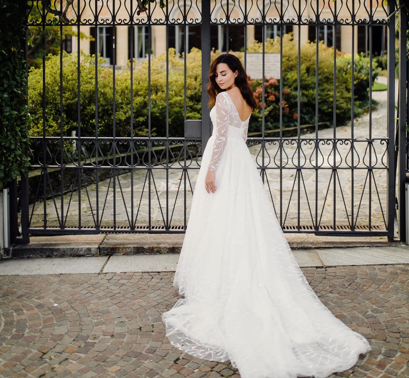A woman in a long white wedding dress stands in front of a wrought iron gate, looking over her shoulder.