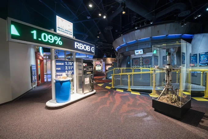 Museum exhibit displays on petroleum, featuring a gas pump, price ticker, blue barrel, and machinery models under bright lighting and informational signage.