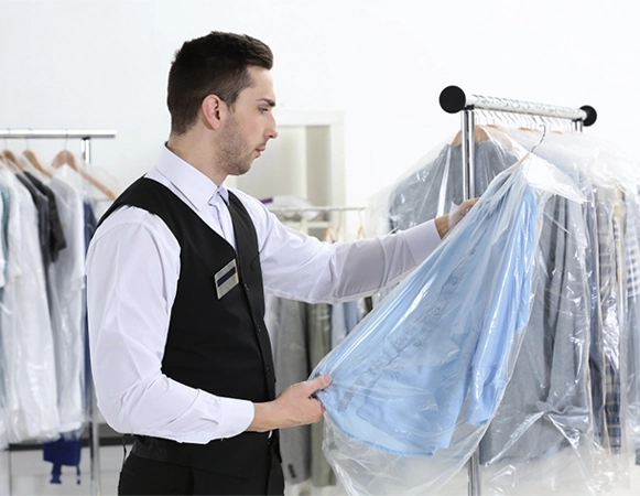 A man in a vest and name tag inspects a light blue shirt covered in plastic on a rack at a dry cleaning facility.