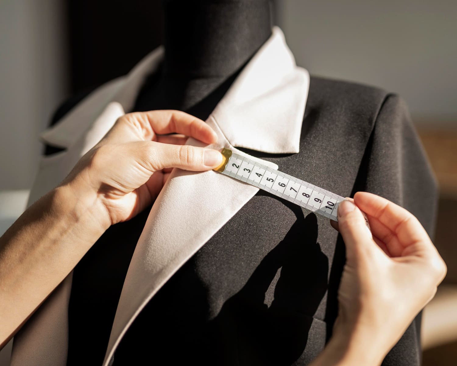 Hands holding a measuring tape against the lapel of a black and white blazer on a dress form, indicating garment fitting or tailoring.