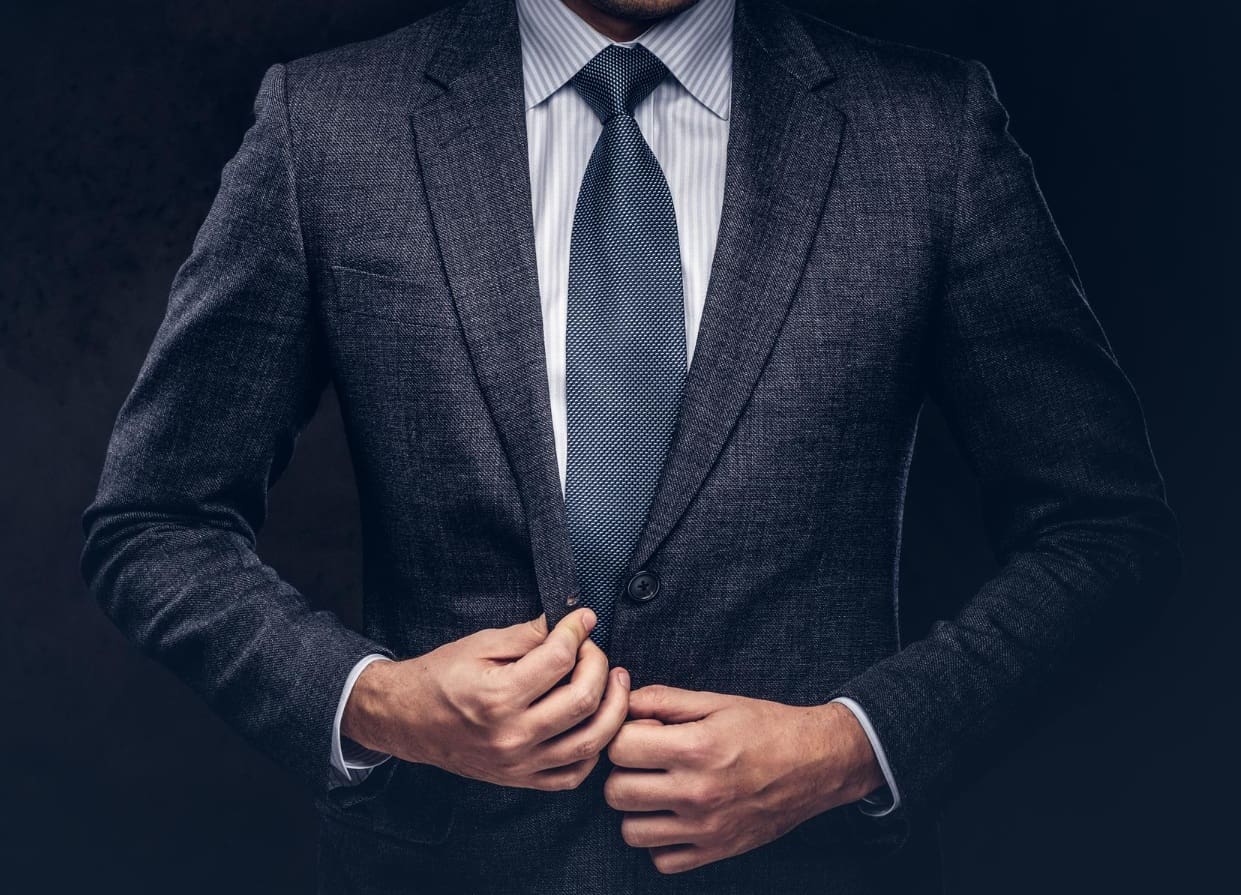 Man in a dark suit and blue tie buttoning his jacket, shown from the shoulders to the waist against a dark background.