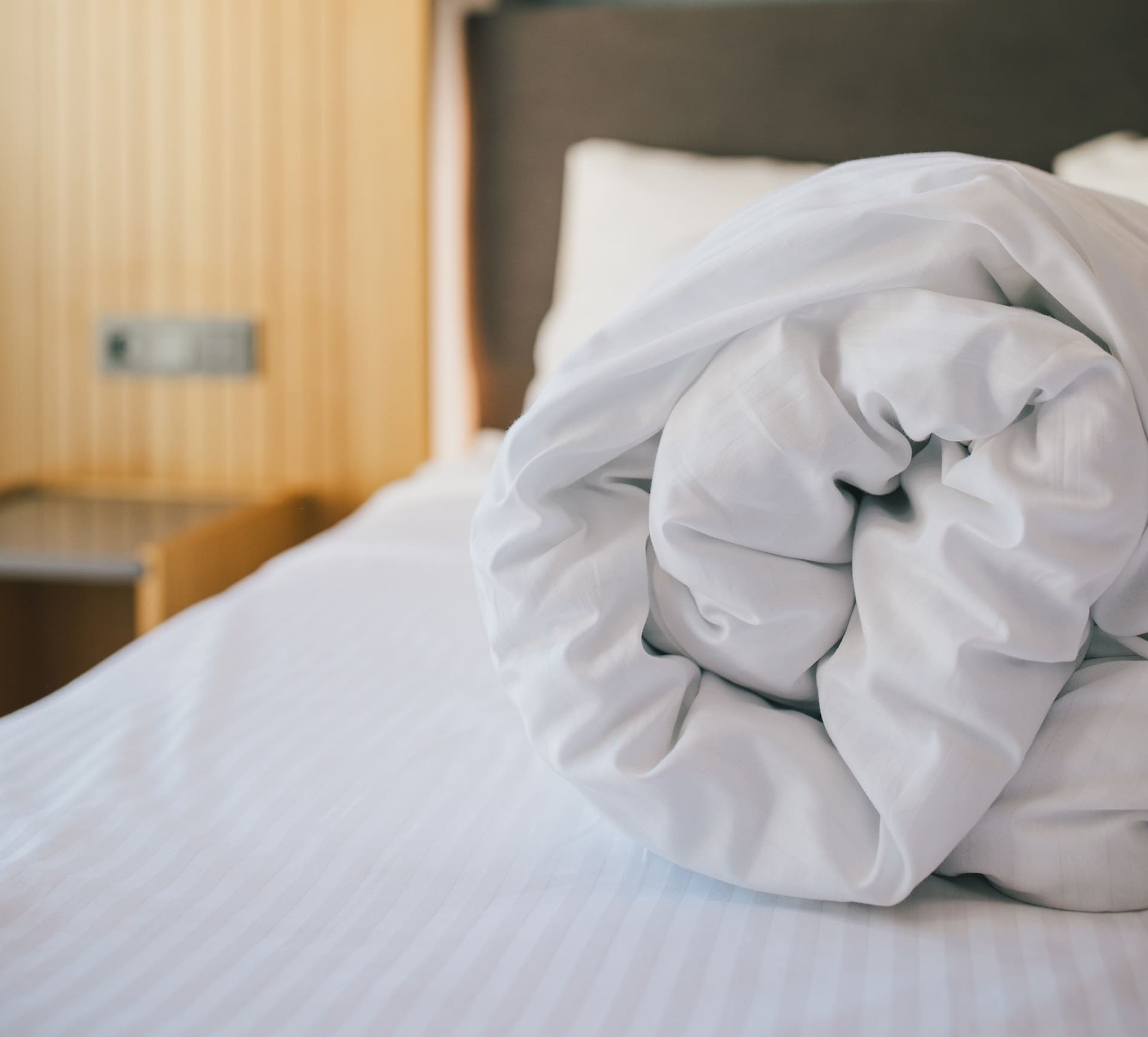 A neatly rolled white comforter lies on a made bed with white sheets in a softly lit bedroom.