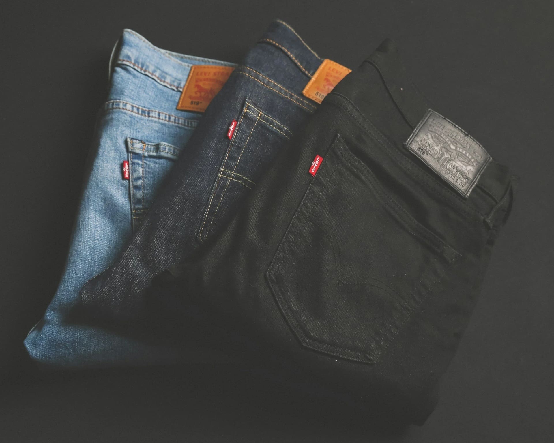 Three pairs of Levi’s jeans, in light blue, dark blue, and black, are folded and stacked side by side against a dark background.