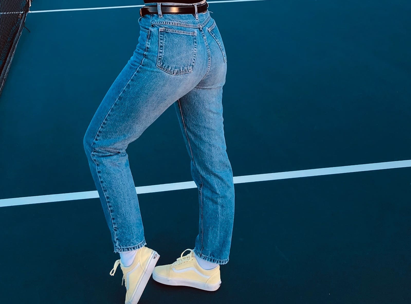 Person standing on a tennis court wearing blue jeans, a black belt, and yellow sneakers; their upper body is not visible.