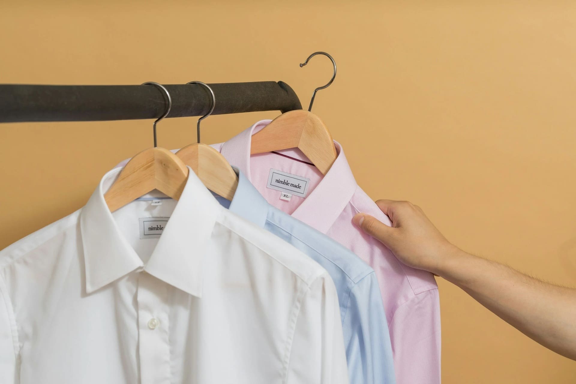 Three collared shirts—white, light blue, and pink—on wooden hangers hanging from a clothing rack, with a hand touching the pink shirt.