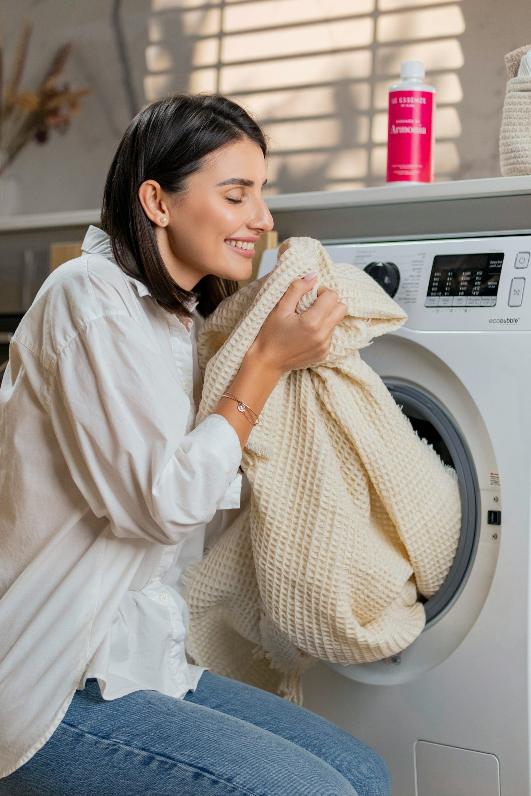 A woman smiles as she holds a beige textured blanket near her face, sitting by a front-load washing machine with a pink bottle of detergent on top.