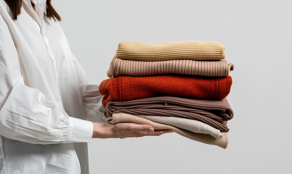 A person in a white shirt holds a neatly folded stack of sweaters in neutral and warm tones against a plain background.