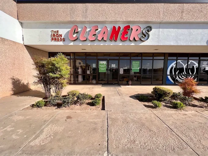 Storefront of a dry cleaners with red and black "CLEANERS" sign above glass doors, sidewalk, and trimmed bushes in front.