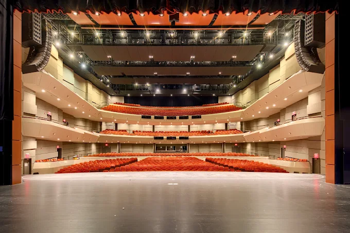 View from the stage of a modern theater auditorium with multiple balconies and rows of red seats, illuminated by ceiling lights.