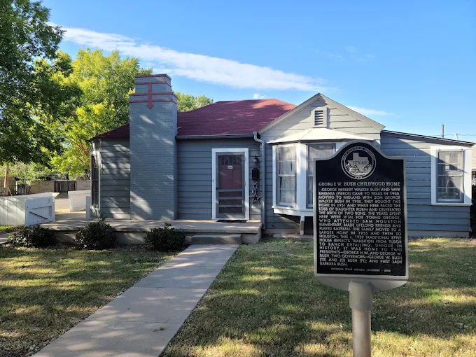 A small gray house with a red roof and a historical marker in front, identifying it as the George H. Bush childhood home, set in a suburban neighborhood with trees and a sidewalk.