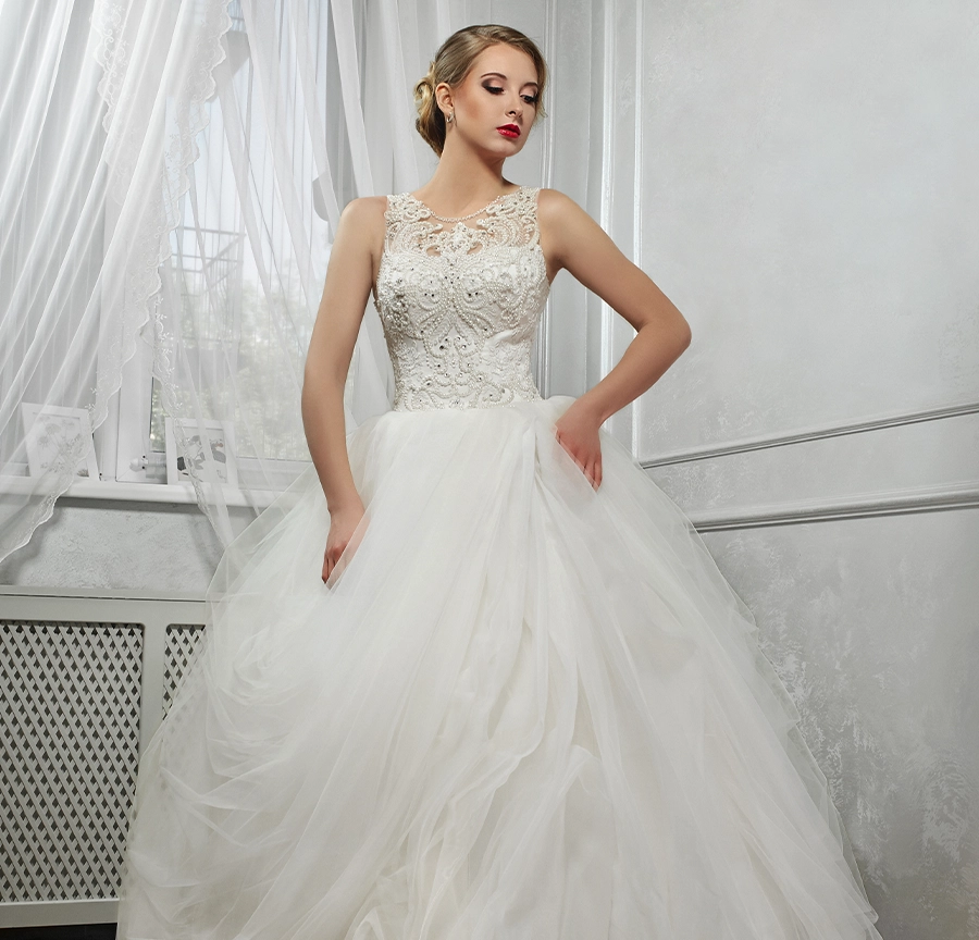 A woman in a sleeveless white lace wedding dress stands indoors by a window with sheer curtains, looking to the side.