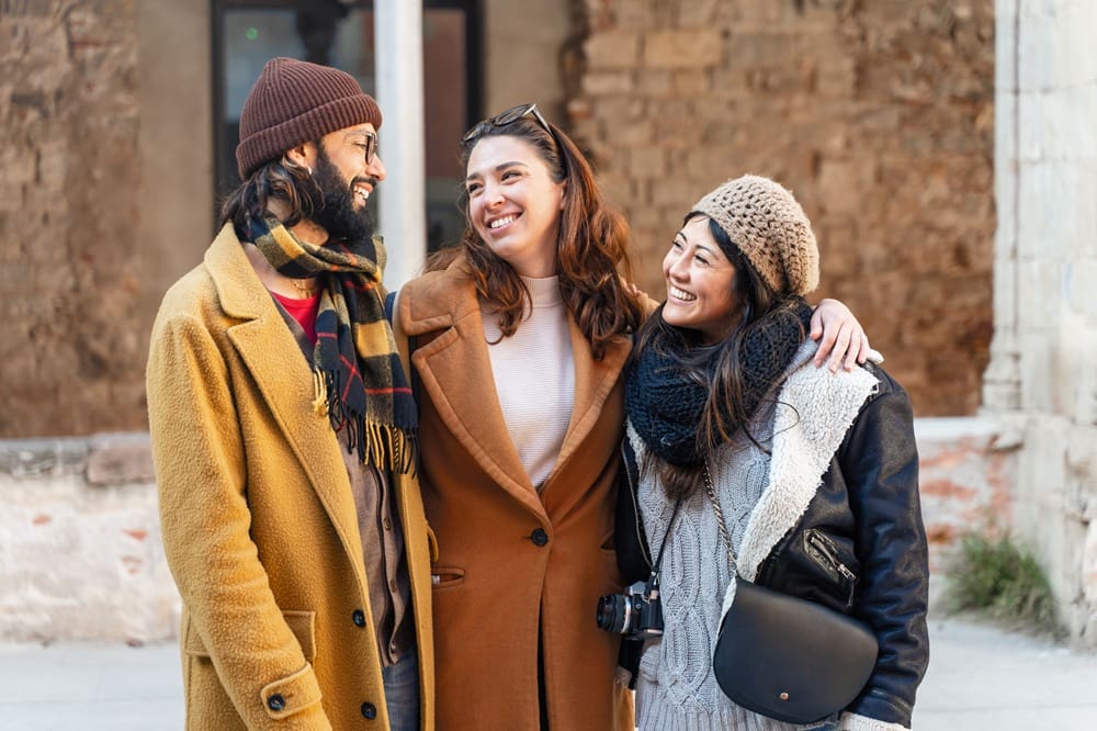 Three people wearing winter coats and hats stand close together outdoors, smiling and talking. One person has a camera hanging around their neck. An old brick wall is in the background.