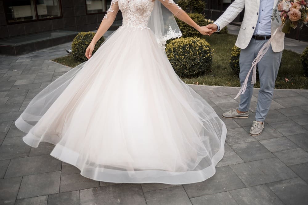 Bride in a white wedding dress and groom in a light jacket and pants holding hands outdoors, with greenery and pavement in the background.