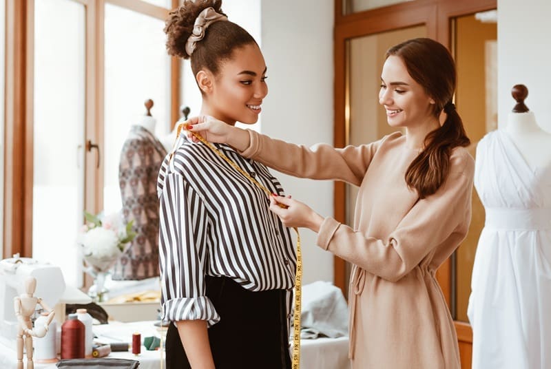 Two women in a fashion studio, one measuring the other's shoulders with a measuring tape. Dresses, fabrics, and a mannequin are visible in the background.