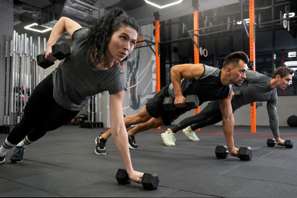 Three people in athletic wear perform renegade rows with dumbbells in a gym with black flooring, orange equipment, and weightlifting bars in the background.