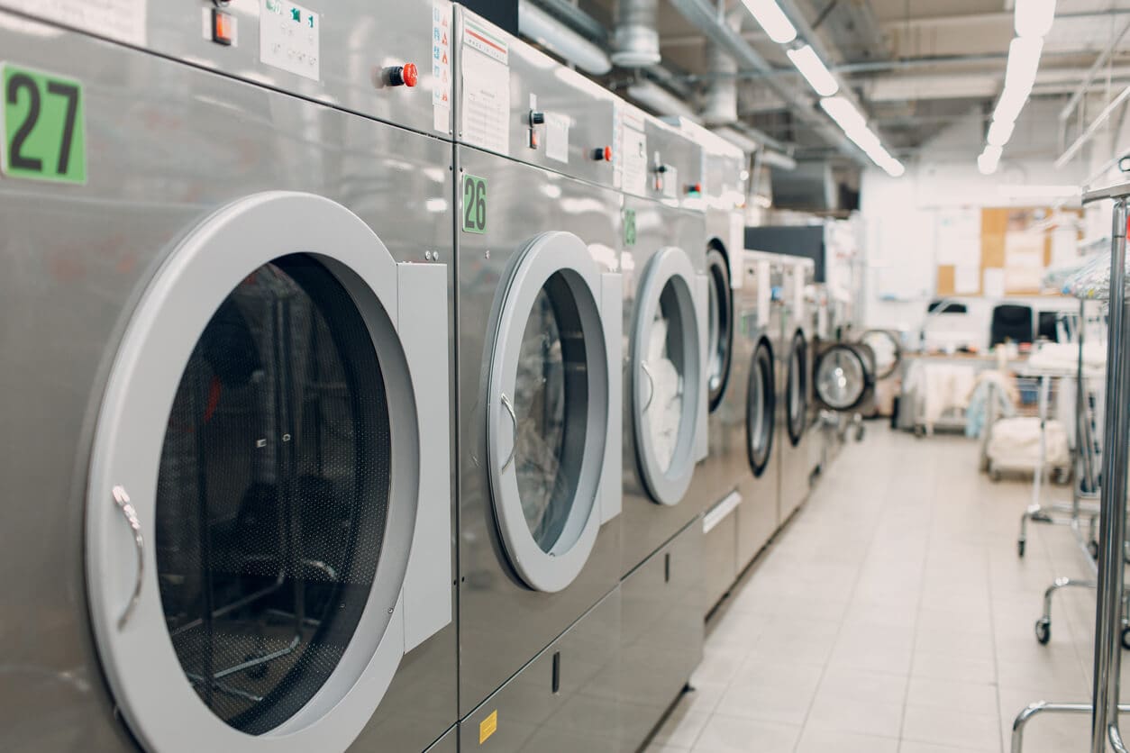 Row of industrial washing machines in a laundromat, with tiled flooring and fluorescent lighting visible.