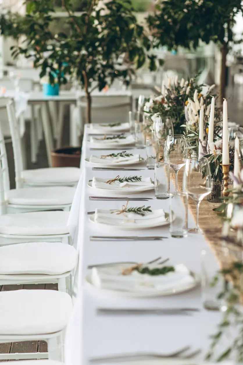 A long table set for a formal event with white linens, plates, glassware, cutlery, candles, and green foliage centerpieces. White chairs line both sides of the table.