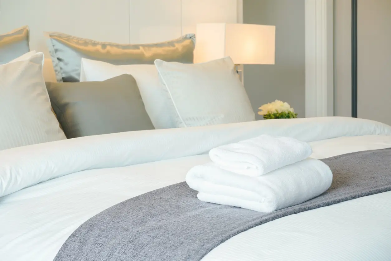 A neatly made bed with white linens, gray accent blanket, several pillows, and two folded white towels placed on top. Lamp and small flower arrangement visible in the background.
