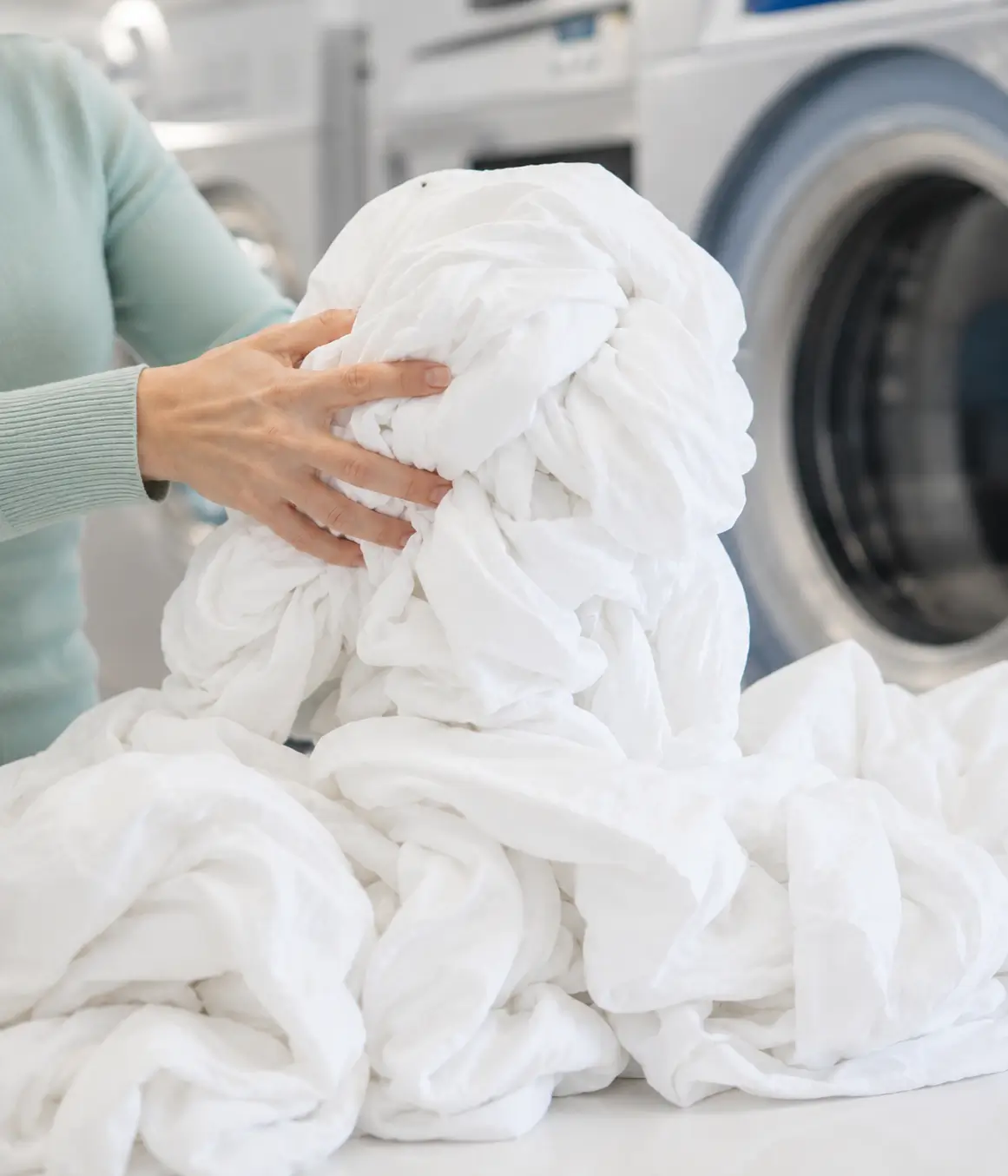 A person holds a large pile of clean white laundry in front of industrial washing machines at a laundromat.